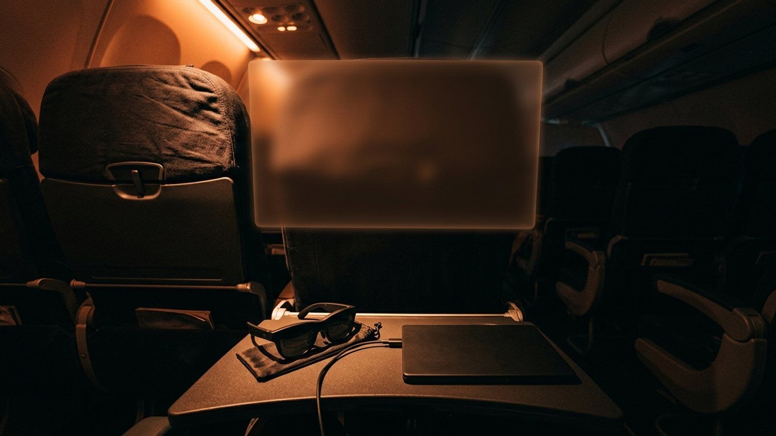 Display glasses cabled to a closed laptop on an airplane tray table, with a large blank virtual screen floating in the air above the seat.