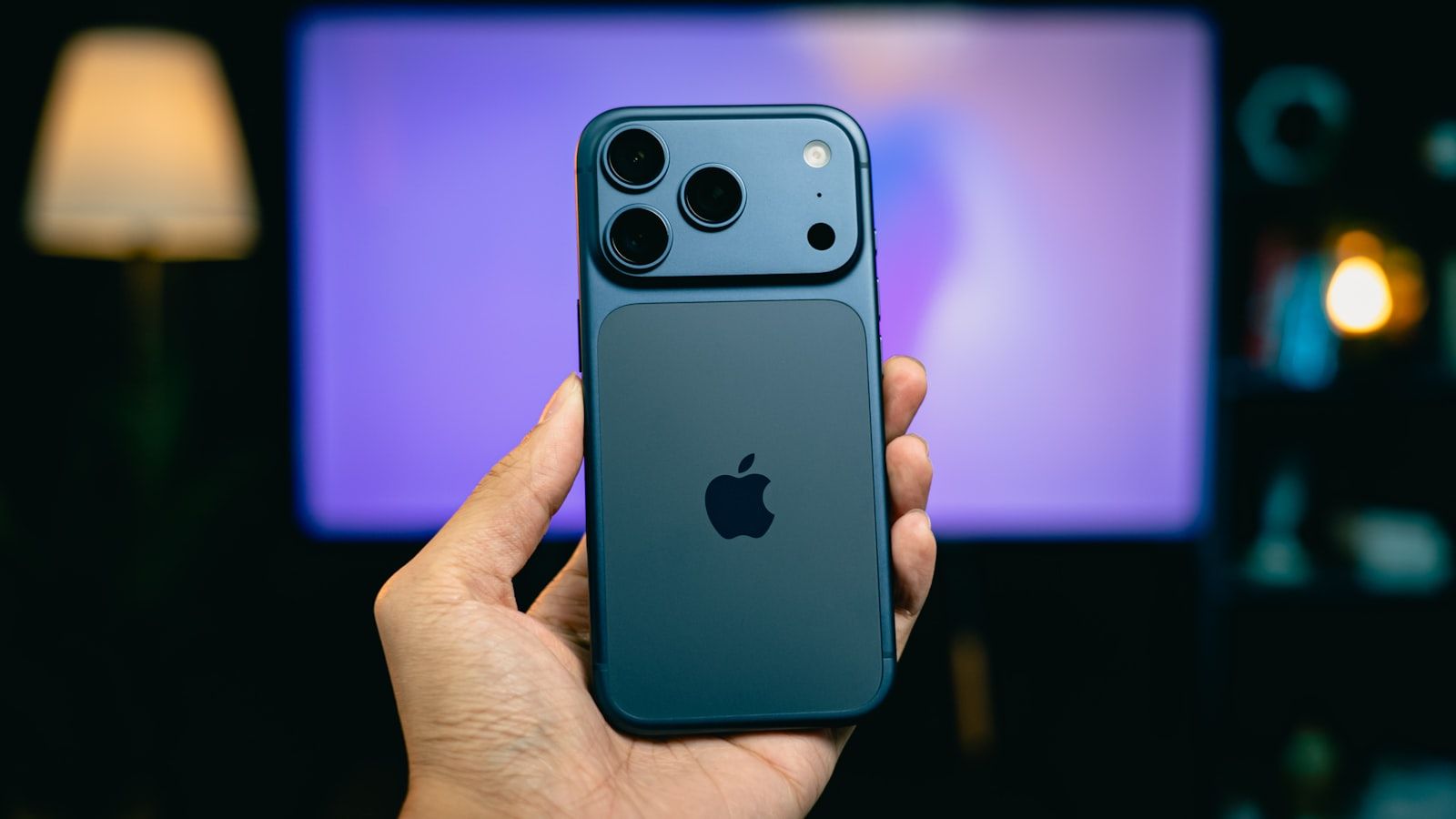 A hand holding a dark blue iPhone Pro showing the Apple logo and rear camera array